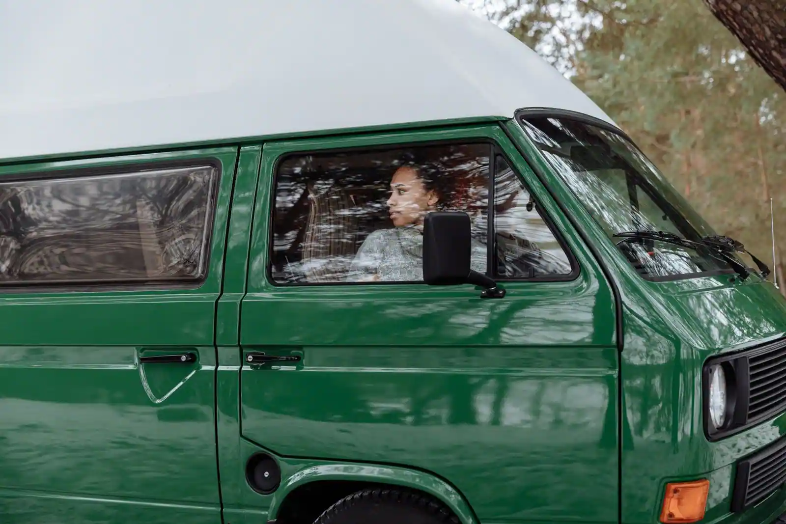 Camping-car aménagé de couleur verte stationné parmi les arbres, une personne installée à l'intérieur.