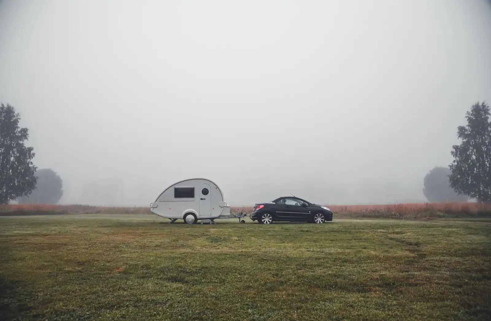 Petite caravane attelée à une voiture, stationnée dans un champ par temps de brume