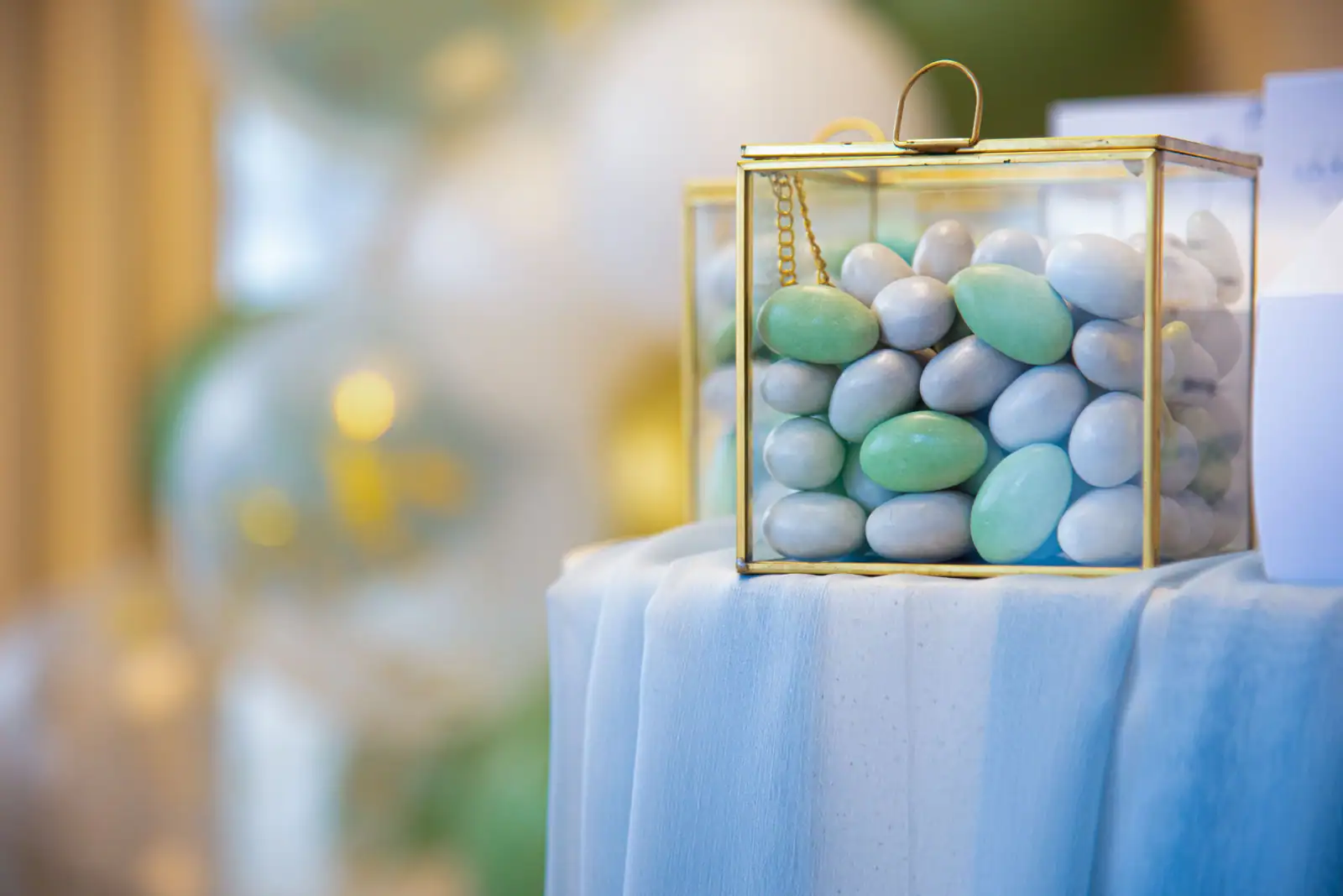 Dragées de baptême pastel vert d'eau, gris et blanc dans une boîte vitrée posée sur un drapé bleu