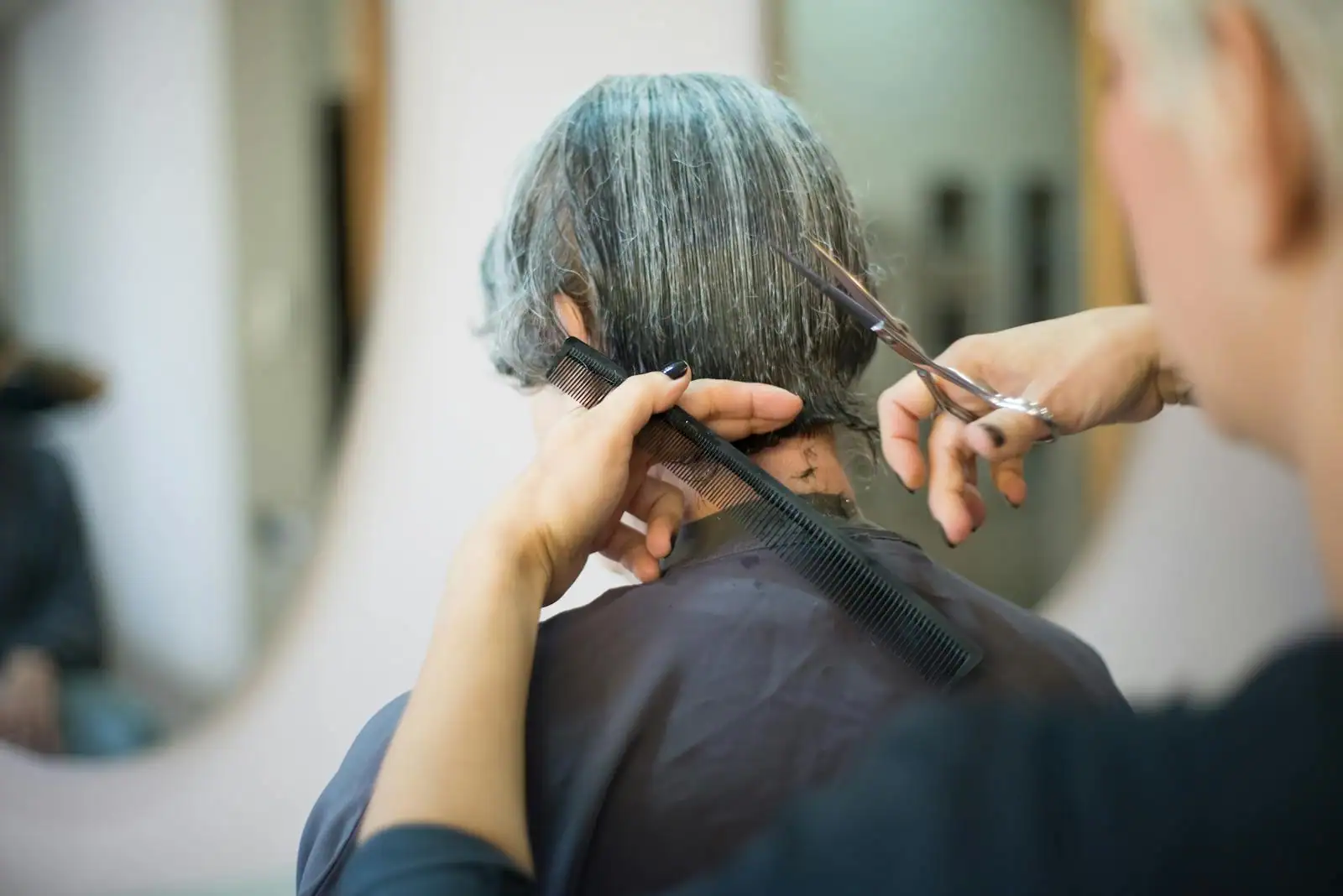 Un coiffeur coupe les cheveux d'une cliente au salon, peigne et ciseaux en main, vue de dos