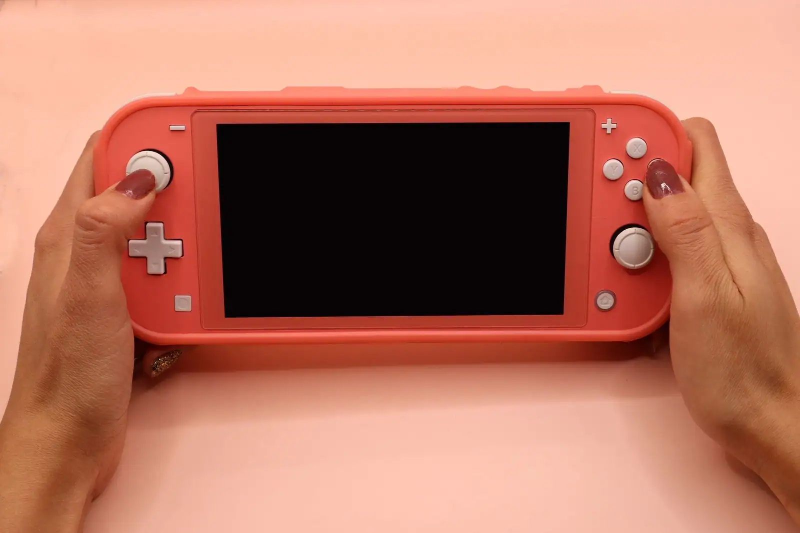 Console portable Nintendo Switch Lite de couleur corail tenue à deux mains sur un fond rose pâle
