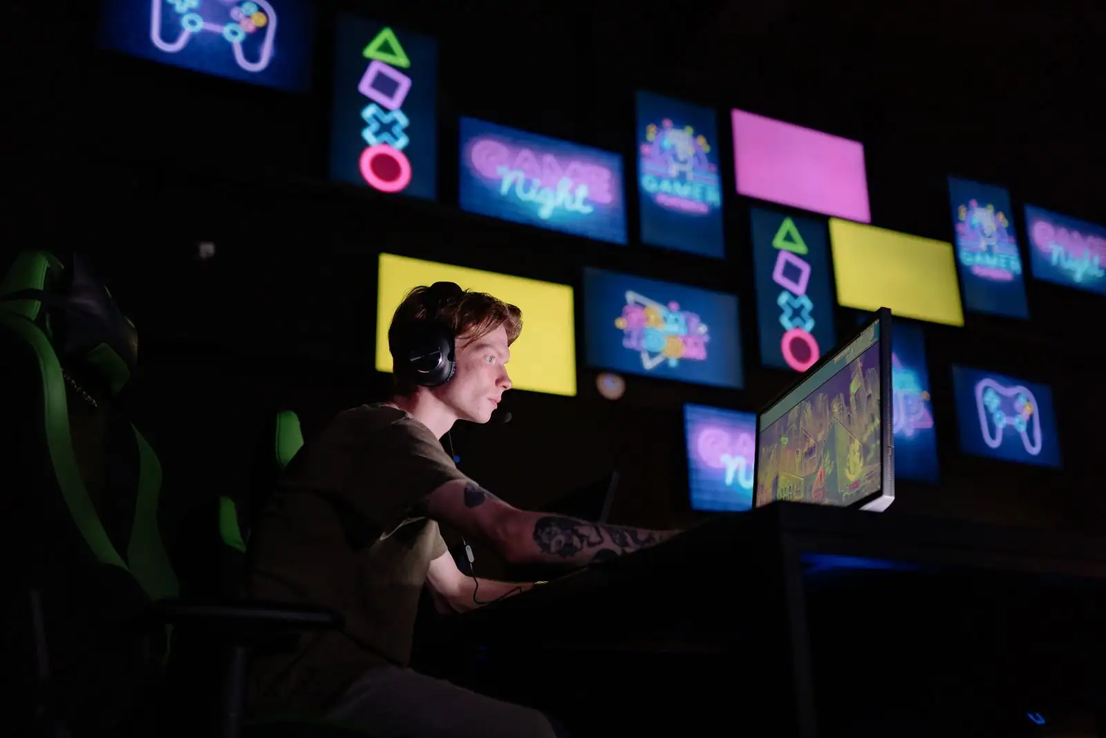 Joueur avec casque, de profil devant un écran de jeu vidéo, dans une pièce sombre éclairée par des enseignes lumineuses de jeux