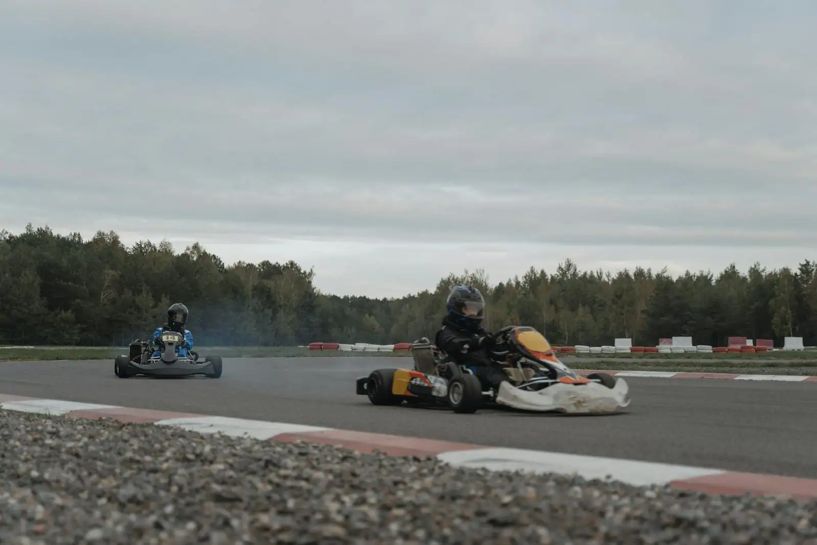 Deux karts sur un circuit asphalté en extérieur, pilotes casqués, bordures rouge et blanc.
