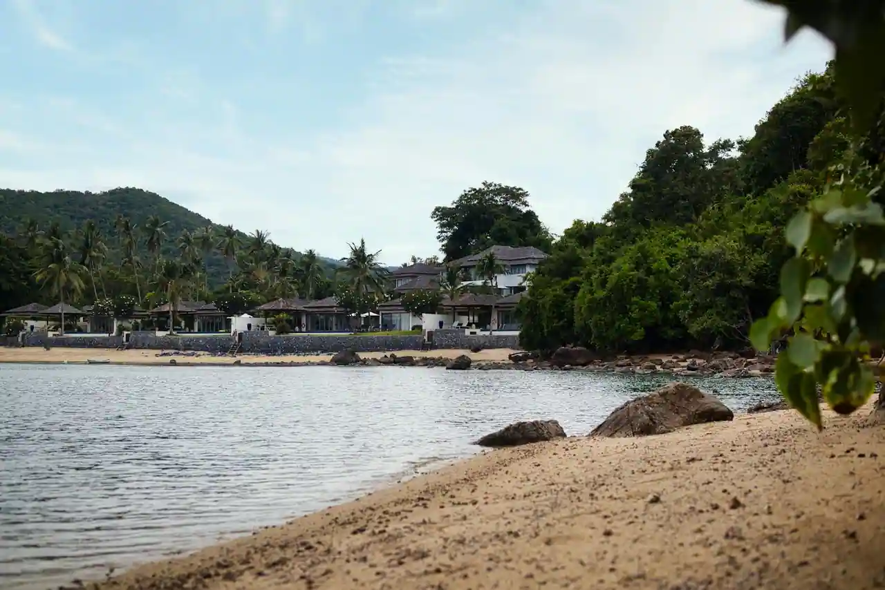 Plage tropicale paisible bordée de villas et de collines verdoyantes sous un ciel bleu.
