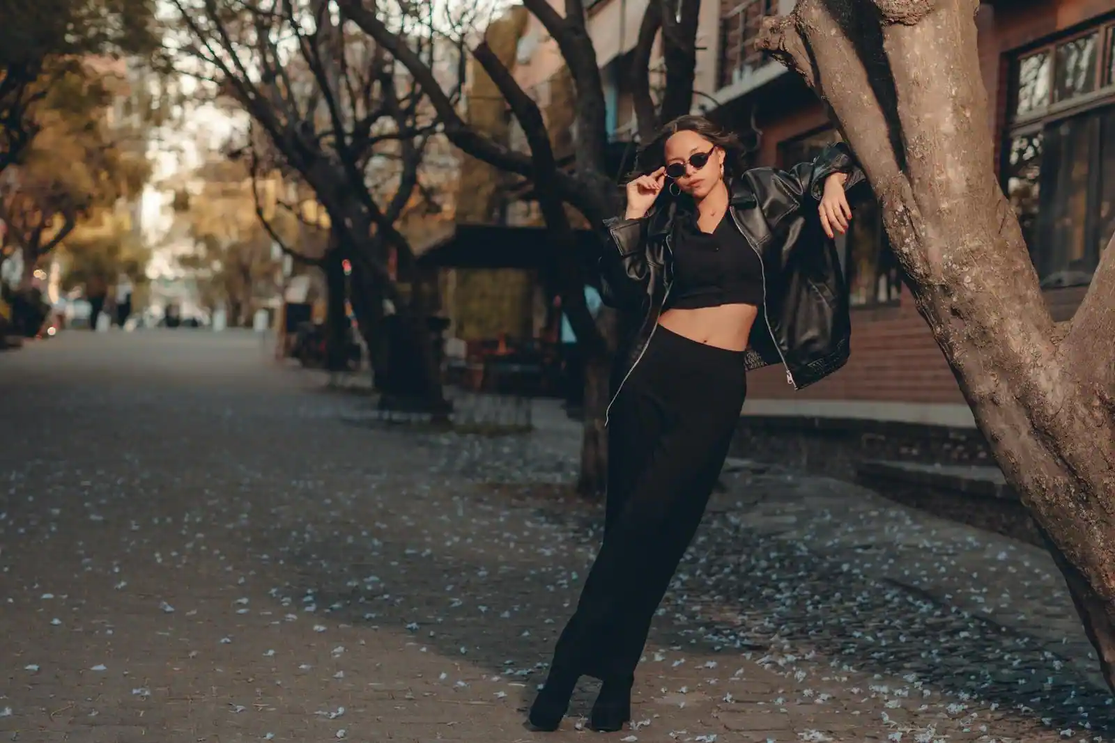 Femme en tenue urbaine noire posant dans une rue arborée, allure mode décontractée et chic.