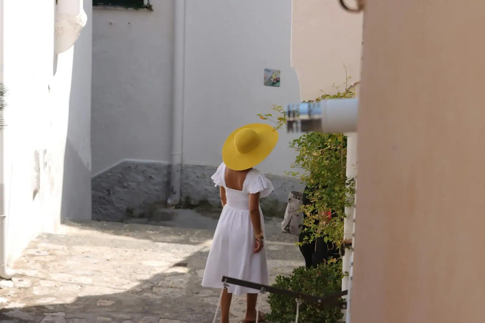 Femme de dos en robe blanche et grand chapeau jaune dans une ruelle italienne ensoleillée.