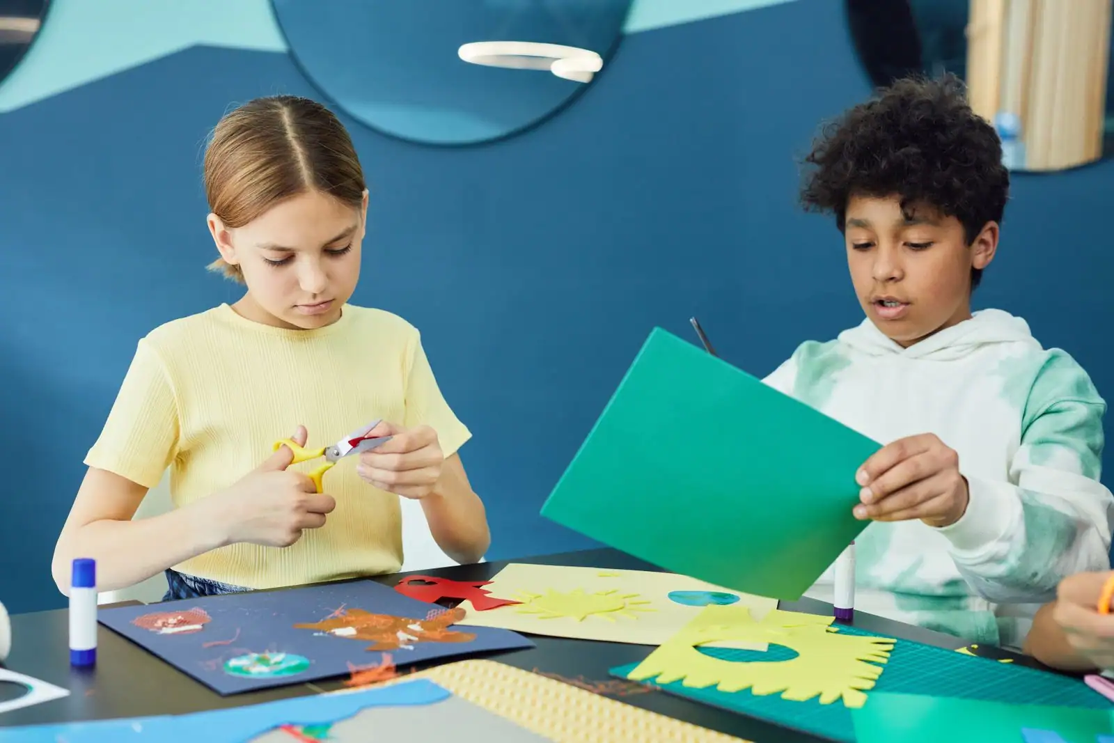 Deux enfants découpent des papiers colorés à table, avec ciseaux, bâton de colle et découpages