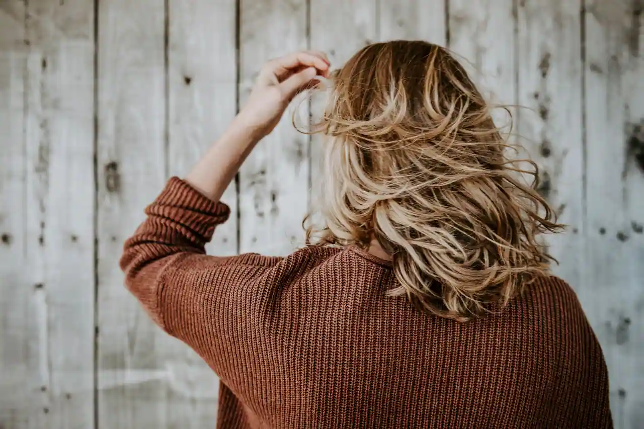 Vue de dos d'une femme aux cheveux blonds mi-longs ondulés, aux longueurs plus claires que les racines