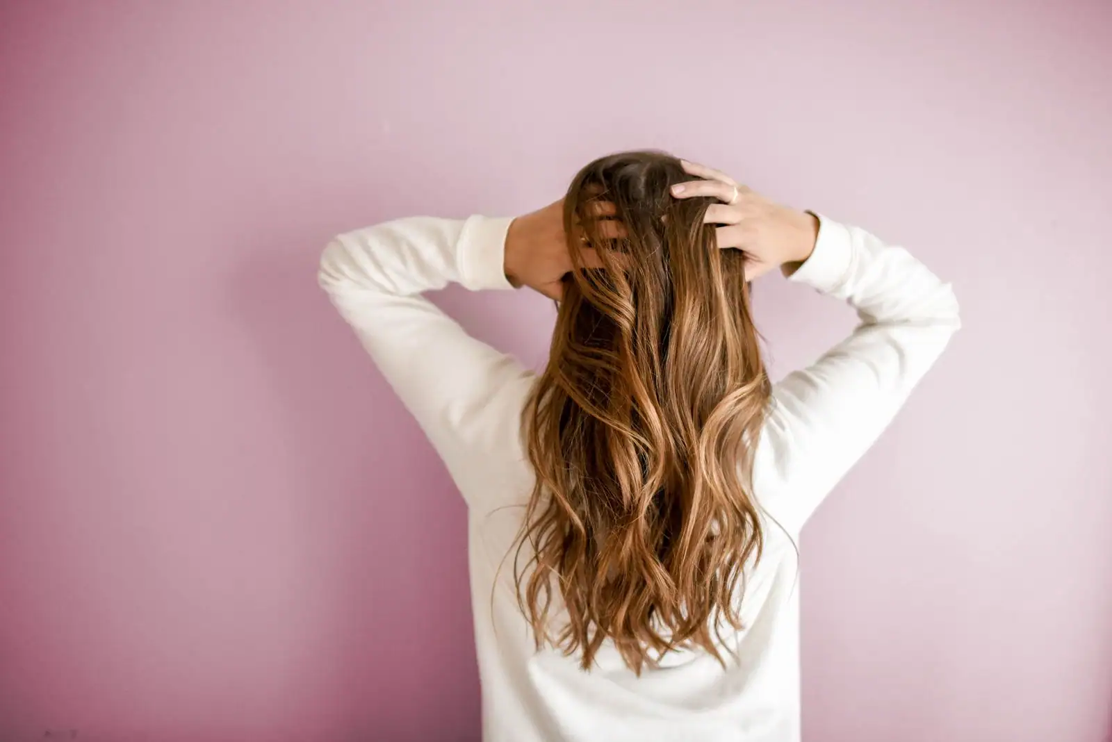 Vue de dos d'une chevelure ondulée brune aux reflets caramel et au balayage doré, sur fond rose.