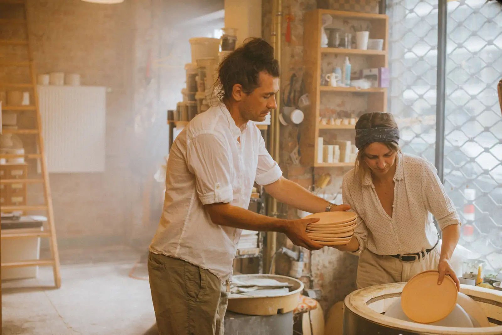 Deux artisans céramistes manipulent des assiettes empilées près du four dans un atelier de poterie