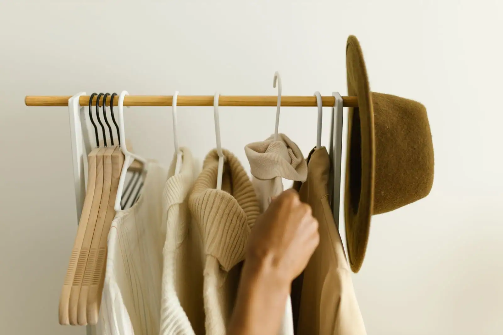 Tringle en bois où sont suspendus un pull, une chemise et un manteau beiges, avec un chapeau en feutre au bout