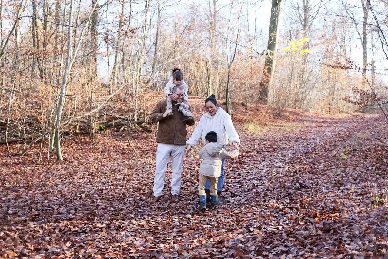 Famille en promenade sur un chemin forestier couvert de feuilles mortes, un enfant juché sur les épaules.