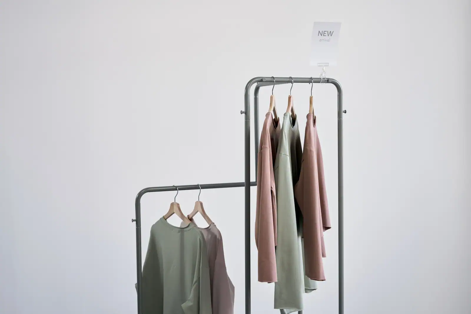 Des blouses neuves rose et vert d'eau suspendues sur des cintres en bois, sous un panneau nouveautés