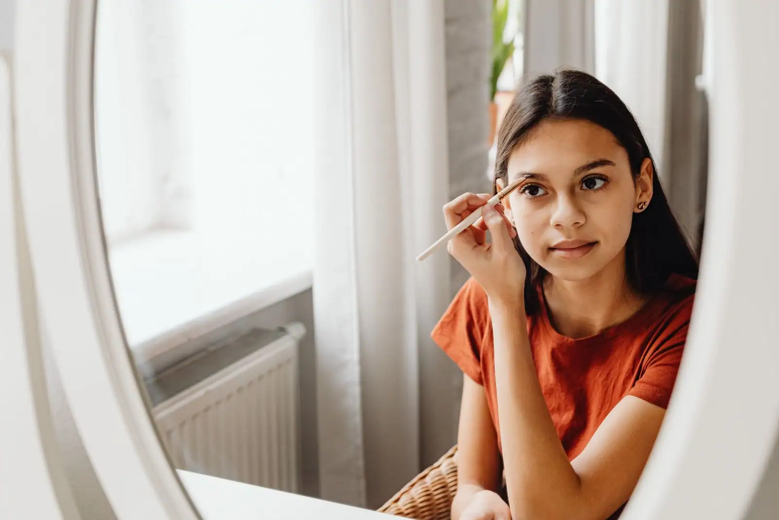 Une jeune fille trace un trait au crayon à sourcils devant son miroir, à la lumière d’une fenêtre.