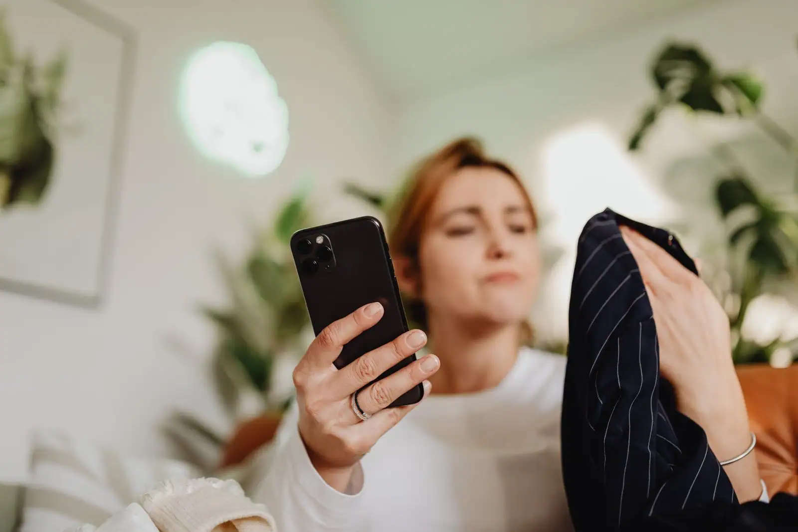 Une femme tient un vêtement rayé et consulte son smartphone à la maison, en plein achat de mode en ligne.