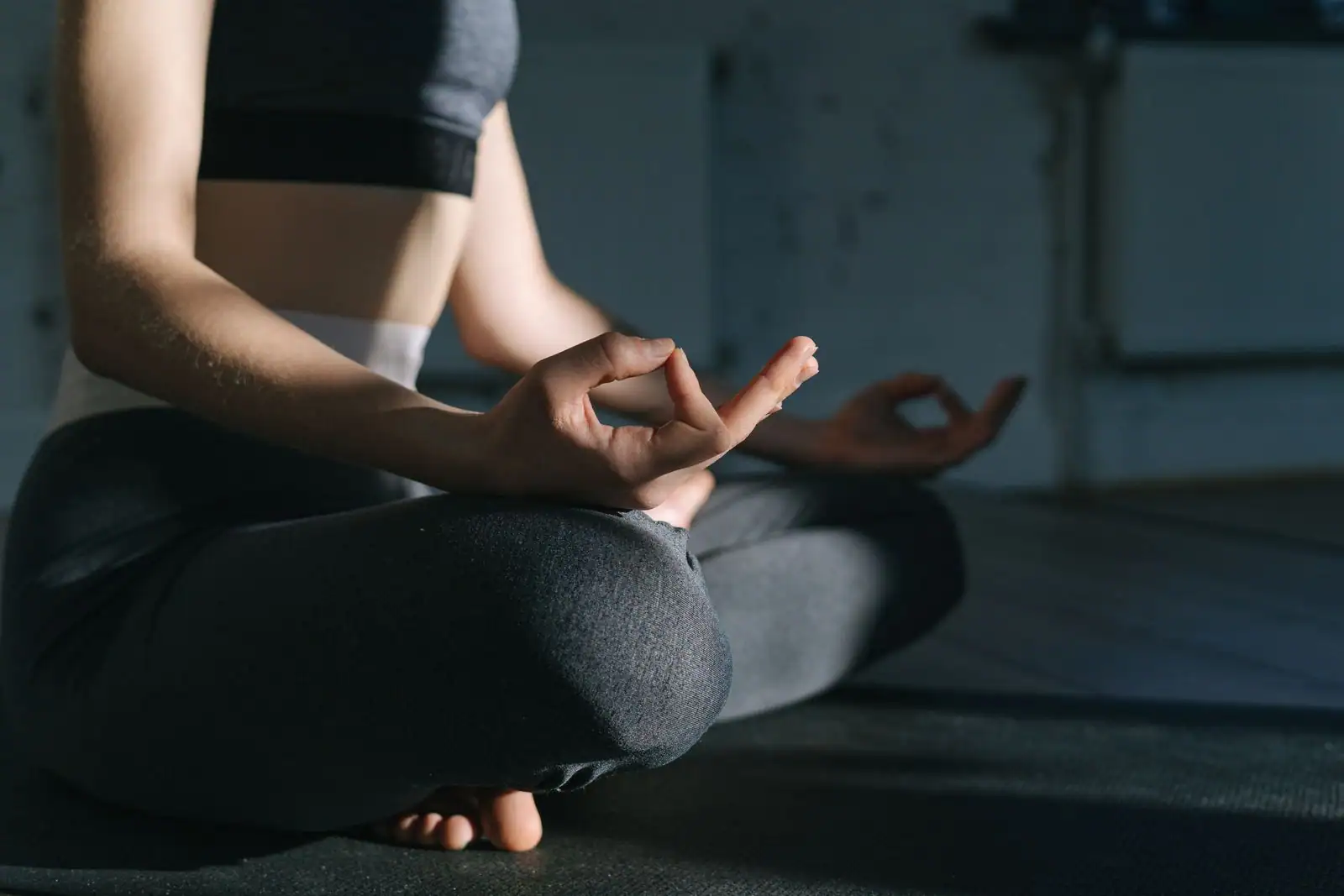 Une personne assise en tailleur sur un tapis, les mains en geste de méditation, dans une pièce sombre