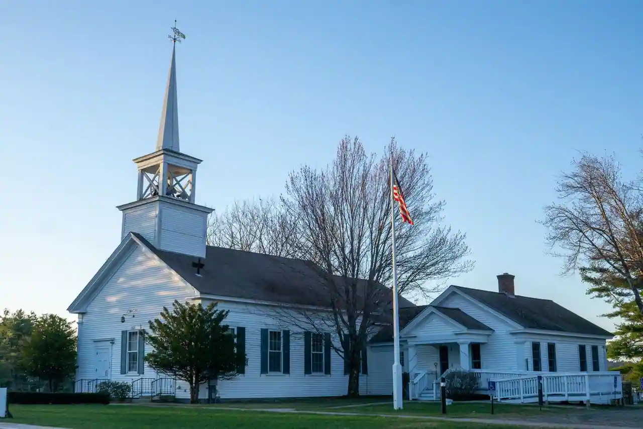 Une église américaine traditionnelle au clocher blanc, drapeau des États-Unis devant, à la lumière du soir.