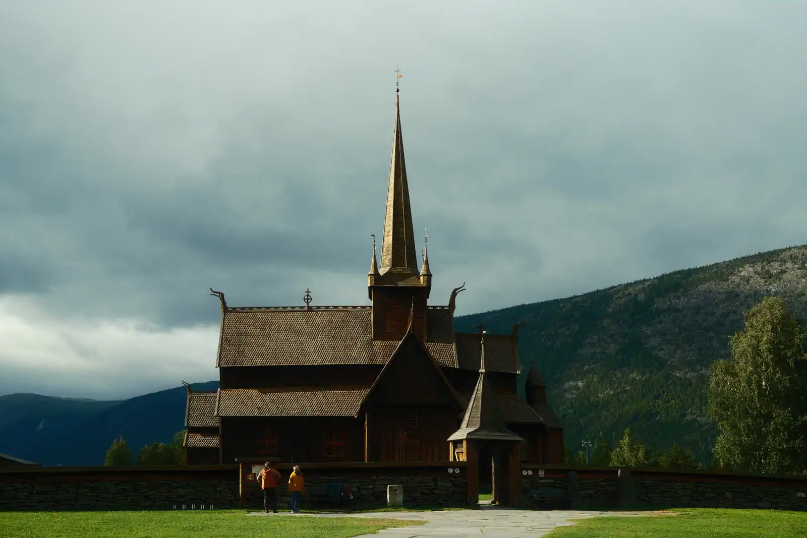 Église en bois debout norvégienne, toits étagés en bardeaux et flèche centrale, montagnes en fond