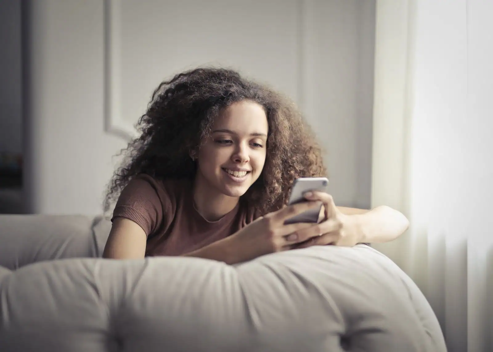 Jeune femme souriante en train d'écrire un message sur son smartphone, installée chez elle.