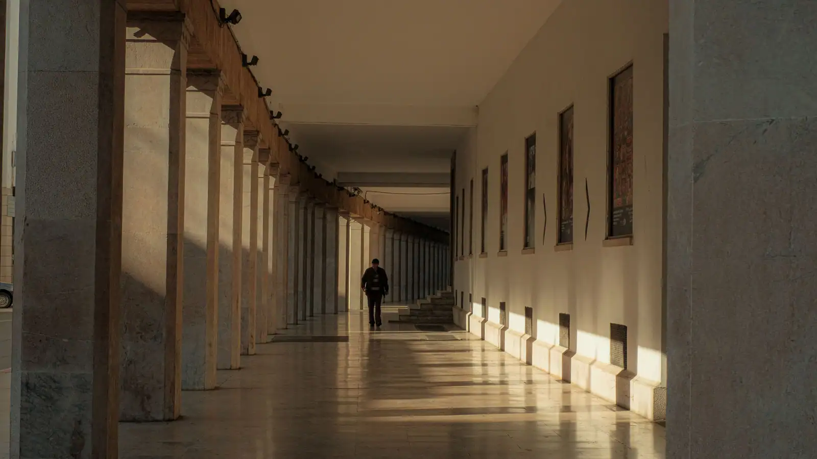 Une silhouette isolée avance dans une longue galerie à colonnes, éclairée par une lumière rasante.