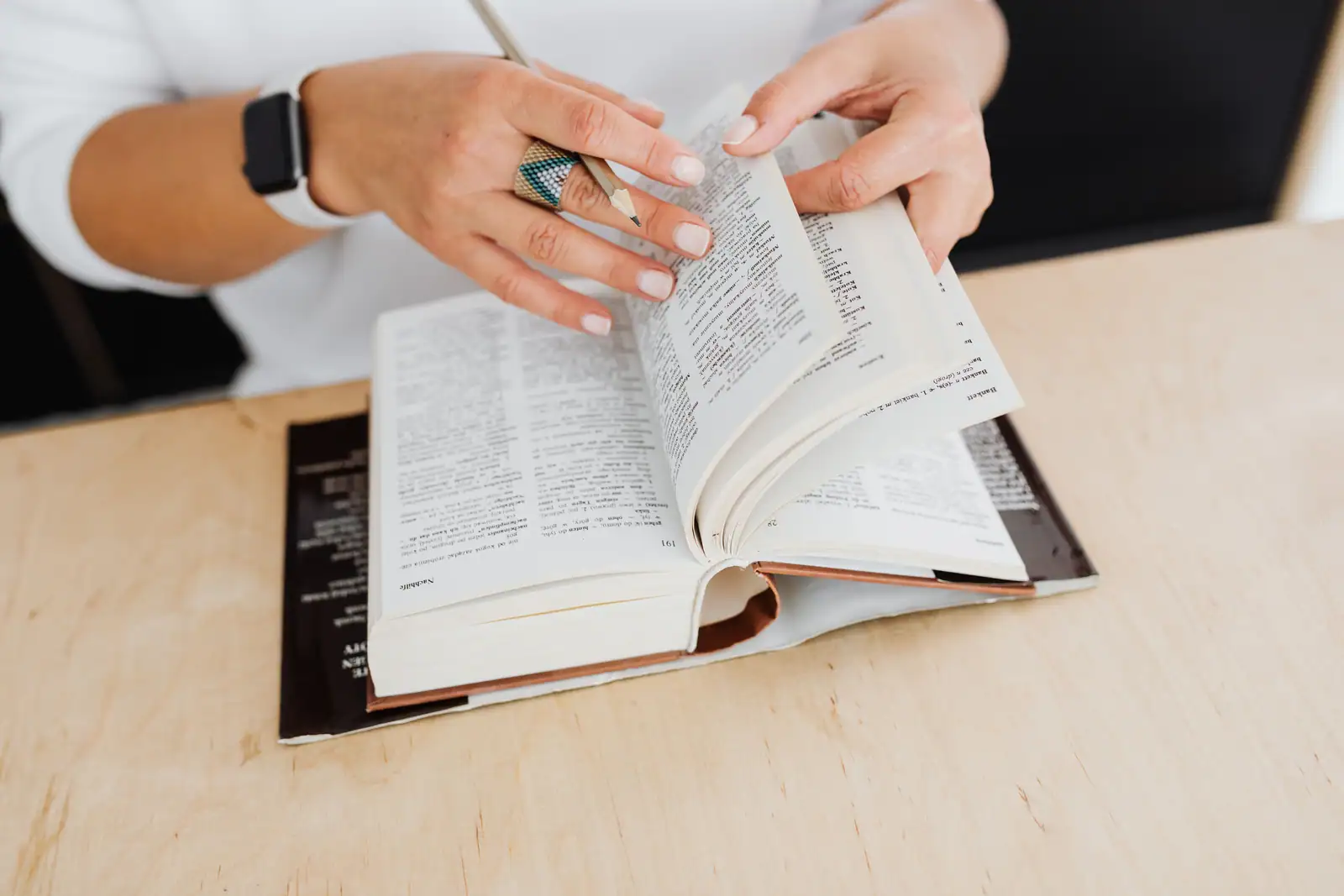 Les mains d’une femme feuillettent un dictionnaire épais posé sur un bureau en bois clair.
