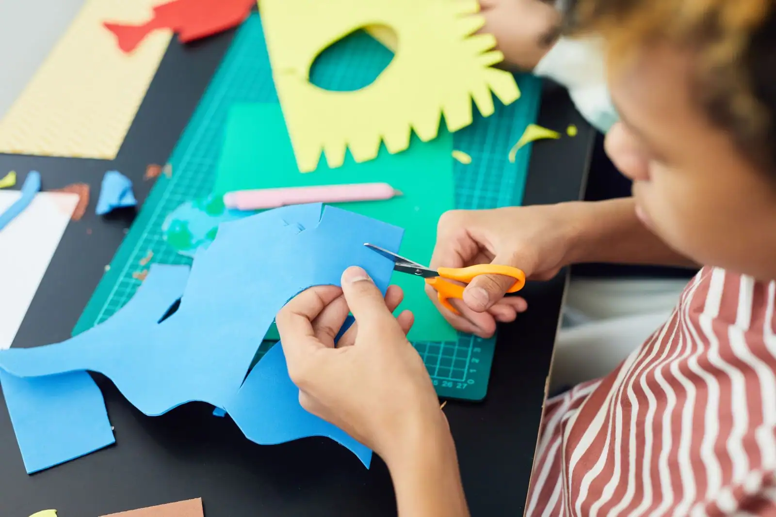 Enfant découpant une forme dans du papier coloré aux ciseaux, sur un tapis de découpe